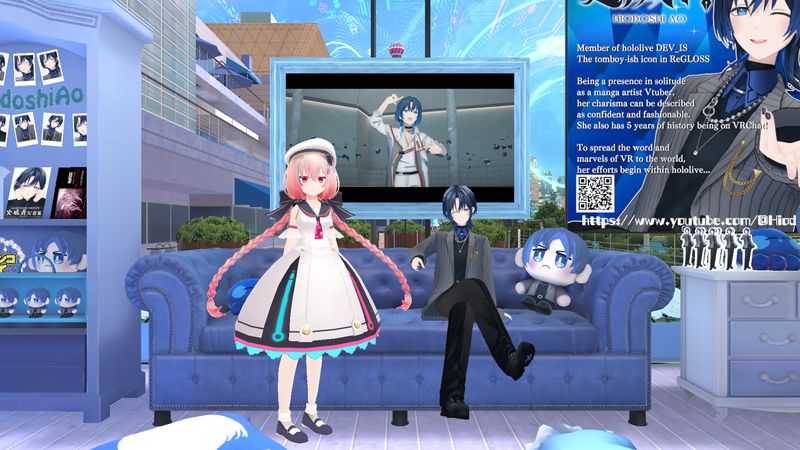 A girl with pink twintails standing beside Hiodoshi Ao—a VTuber with short dark blue hair in a black suit—sitting on a blue sofa along with a stuffed toy version of herself. Various pictures and merchandise of her are on the side, while a music video from ReGLOSS plays in the booth’s background.